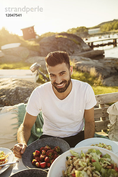 Porträt eines glücklichen jungen Mannes beim Mittagessen am Steg bei Sonnenschein
