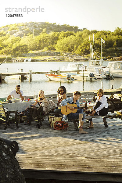 Freunde  die dem Mann beim Gitarrespielen während der Party im Hafen an einem sonnigen Tag zuhören.