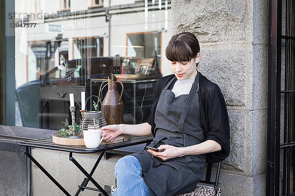 Junge Besitzerin mit Smartphone beim Sitzen mit Kaffeetasse am Tisch gegen den Laden
