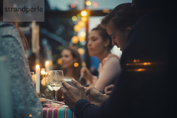 Abgeschnittenes Bild eines jungen Mannes  der während der Dinnerparty inmitten von Freunden am Esstisch sitzt.