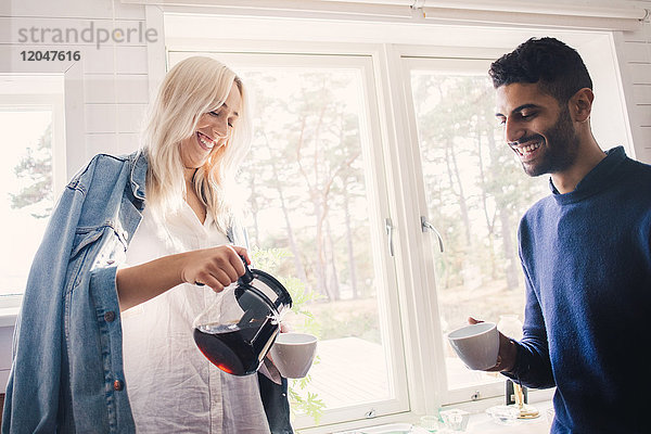 Lächelnde junge Frau  die Kaffee in die Tasse gießt  während sie zu Hause bei einem Freund steht.