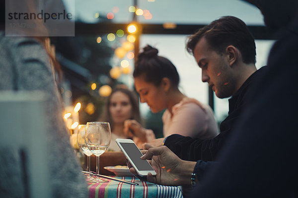 Beschnittenes Bild eines Mannes  der ein Smartphone benutzt  während er unter Freunden am Esstisch sitzt.