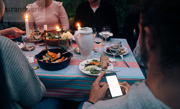Beschnittenes Bild eines jungen Mannes  der ein Smartphone benutzt  während er mit Freunden zu Abend isst.