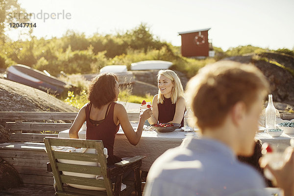 Lächelnde junge Freundinnen sitzen am Tisch und genießen das Sommeressen an einem sonnigen Tag.
