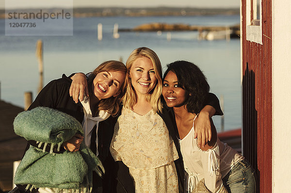 Porträt von lächelnden jungen Freundinnen  die mit den Armen am Hafen gegen den Himmel stehen.