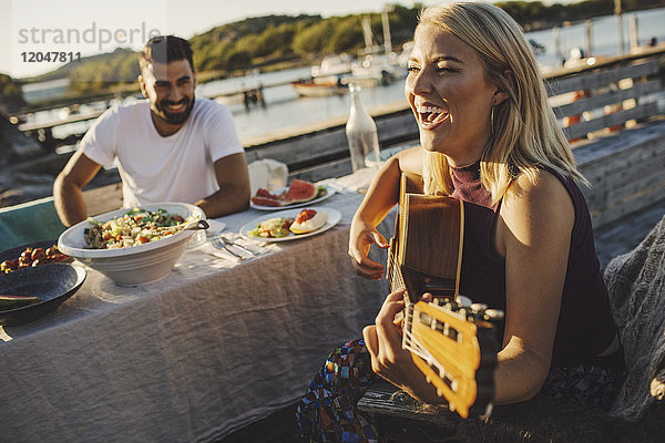 Fröhliche Frau spielt Gitarre  während ein Freund an einem sonnigen Tag am Picknicktisch sitzt.