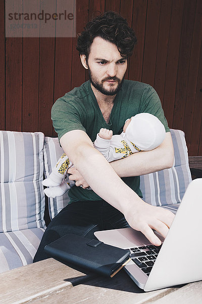 Vater mit Baby mit Laptop im Sitzen auf dem Stuhl an der Wand