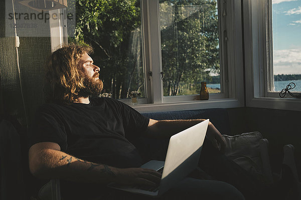 Mann hält Laptop beim Ausruhen am Fenster in der Ferienvilla