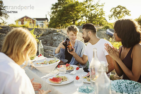 Männer rösten Bierflaschen  während sie mit Freunden am Picknicktisch sitzen.