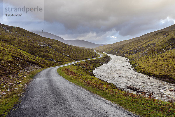 Kurvenreiche Straße mit Fluss und bewölktem Himmel in den Highlands bei Glen Coe in Schottland  Vereinigtes Königreich