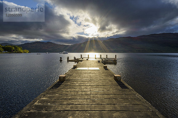 Holzsteg am See mit dramatischen Wolken bei Sonnenaufgang am Loch Lomond in Schottland  Vereinigtes Königreich