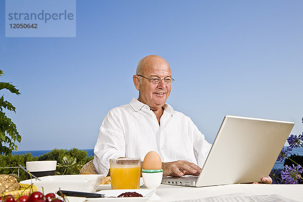 Mann auf der Terrasse  der frühstückt und einen Laptop benutzt