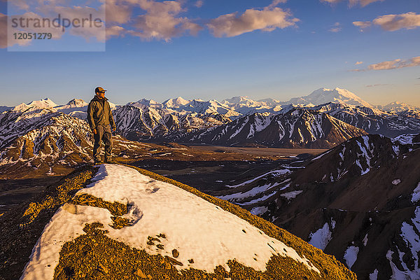 Ein Mann auf einem schneebedeckten Bergrücken in der Nähe des Polychrome Mountain im Denali National Park sieht dem Sonnenuntergang entgegen  mit dem Denali im Hintergrund; Alaska  Vereinigte Staaten von Amerika