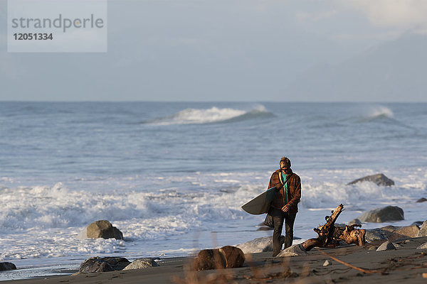 Surfer  der mit einem Surfbrett entlang der Küste in der Nähe von Yakutat  Südost-Alaska  USA  wandert