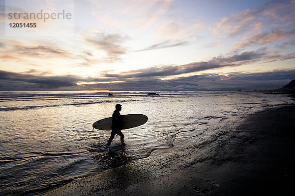 Silhouette eines Surfers  der ein Surfbrett bei Sonnenuntergang zurück zum Ufer trägt; Alaska  Vereinigte Staaten von Amerika