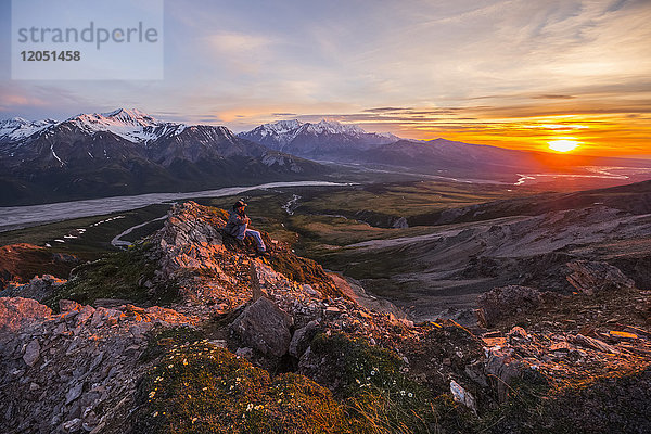 Ein Mann beobachtet einen ruhigen Sonnenuntergang von einem alpinen Sitzplatz hoch über dem Delta River in der Alaska Range; Alaska  Vereinigte Staaten von Amerika