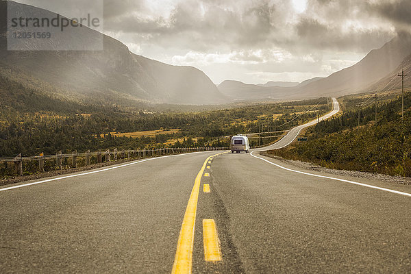 Ein Fahrzeug mit Anhänger fährt eine Straße hinunter  die durch Felder und Berge unter einem bewölkten Himmel führt; Neufundland  Kanada