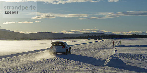 Ein Auto fährt über eine schneebedeckte Straße unter einem blauen Himmel mit Wolken und Bergen in der Ferne; Arjeplog  Provinz Norrbotten  Schweden