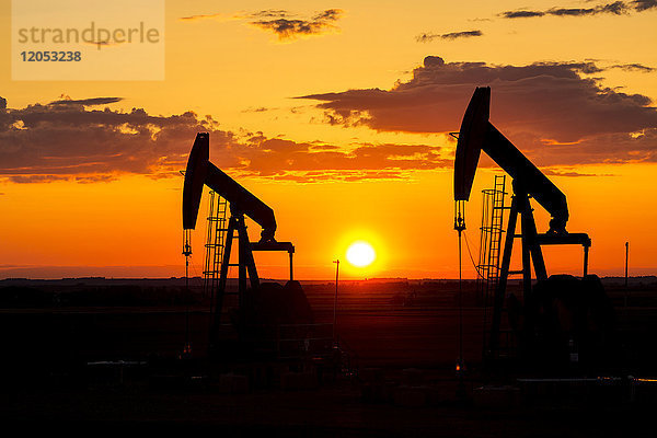 Silhouette von zwei Pumpjacks bei Sonnenaufgang mit einer bunten Orange Sonne  Wolken und Himmel; Alberta  Kanada