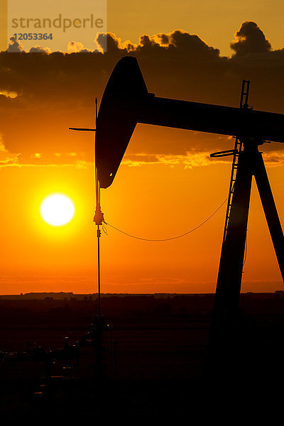 Silhouette eines Pumpjack bei Sonnenaufgang mit einer bunten orangefarbenen Sonne  Wolken und Himmel; Alberta  Kanada