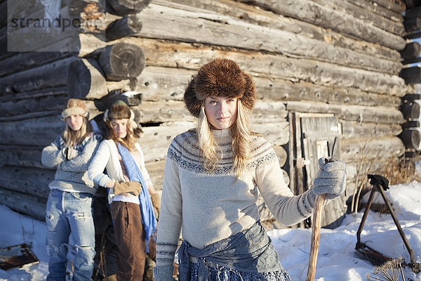 Drei junge Frauen mit Pelzmützen stehen im Schnee mit einer Blockhütte im Hintergrund; Homer  Alaska  Vereinigte Staaten von Amerika