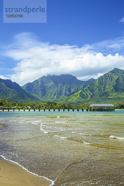 Ein Steg  der zum Meer hinausführt  entlang der Küste  mit Wasser  das auf den Strand gespült wird  im Vordergrund und zerklüfteten  grünen Bergen im Hintergrund; Hanalei  Kauai  Hawaii  Vereinigte Staaten von Amerika