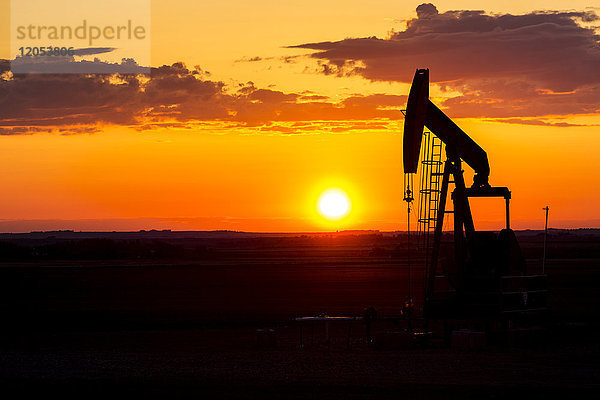 Silhouette eines Pumpjack bei Sonnenaufgang mit einer bunten orangefarbenen Sonne  Wolken und Himmel; Alberta  Kanada