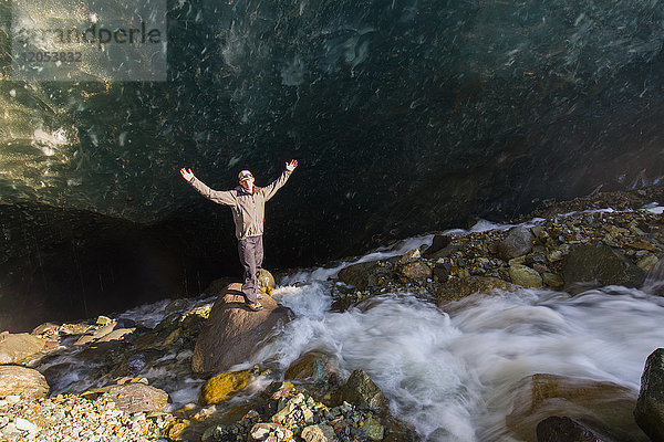 Ein Mann posiert auf einem Felsblock mit ausgestreckten Armen vor einer Höhle unter dem Eis des Root Glacier im Wrangell-St. Elias National Park; Alaska  Vereinigte Staaten von Amerika