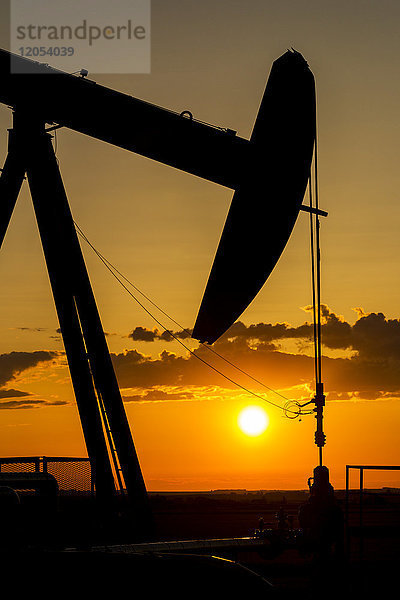 Silhouette eines Pumpjack bei Sonnenaufgang mit einer bunten orangefarbenen Sonne  Wolken und Himmel; Alberta  Kanada