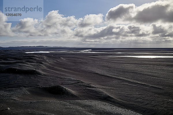 Ein Fluss fließt durch Flussbett aus schwarzem Sand mit Bergen in der Ferne; Island