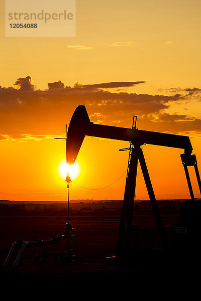 Silhouette eines Pumpjack bei Sonnenaufgang mit einer bunten orangefarbenen Sonne  Wolken und Himmel; Alberta  Kanada