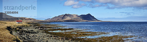 Landschaft entlang der Küste mit Berggipfeln in der Ferne und einem kleinen Gebäude mit rotem Dach entlang der Wasserkante  Halbinsel Snaefellsnes; Island