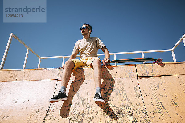 Junger Mann mit Ohrstöpseln und Longboard auf der Halfpipe im Skatepark sitzend