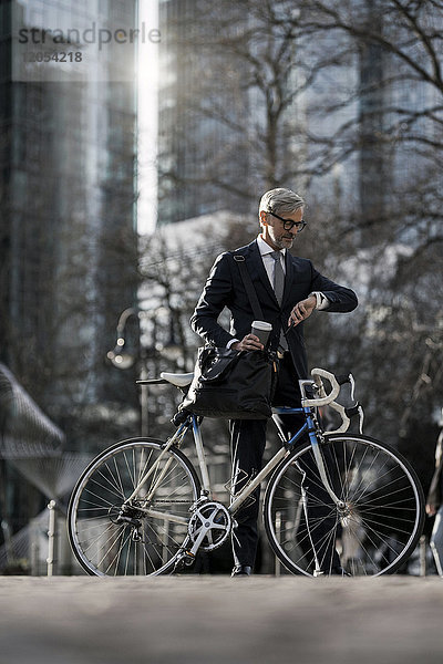 Grauhaariger Geschäftsmann mit Fahrrad und Kaffee zum Mitnehmen in der Stadt mit Blick auf smartwatch