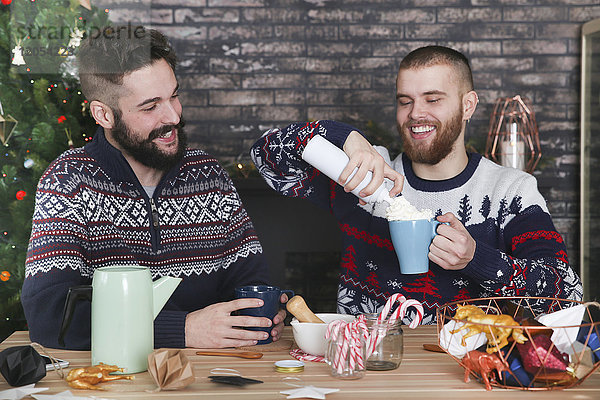 Schwules Paar bereitet heiße Schokolade mit Sahne und gehackten Zuckerstangen zur Weihnachtszeit zu.