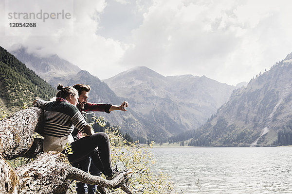 Österreich  Tirol  Alpen  Paar auf Baumstamm am Bergsee sitzend  mit Selfie