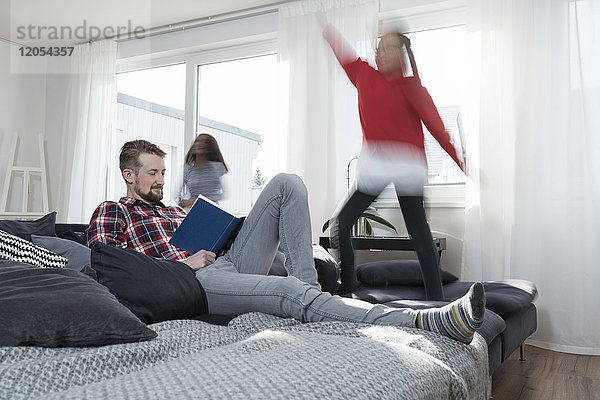 Mädchen springen auf dem Sofa im Wohnzimmer  während Vater ein Buch liest.