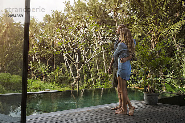 Umarmendes Paar  das auf einer Holzterrasse vor dem Pool steht und eine atemberaubende Aussicht in einem üppigen tropischen Garten genießt.