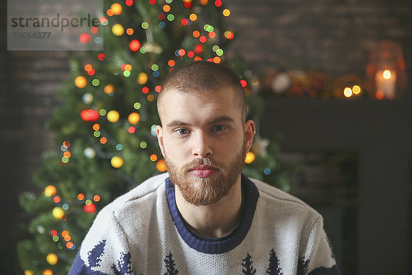 Porträt eines ernsthaften jungen Mannes mit Vollbart vor dem beleuchteten Weihnachtsbaum zu Hause
