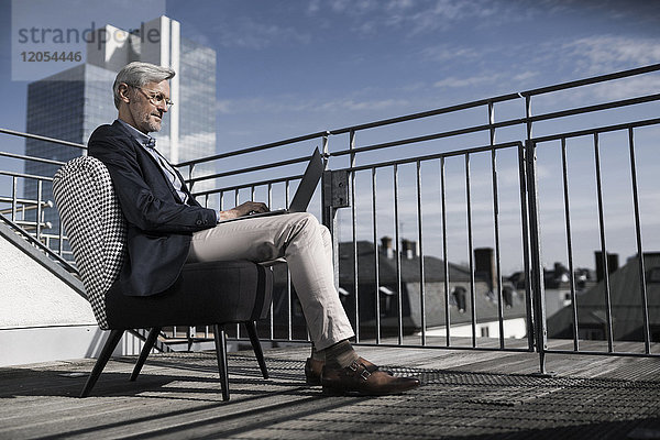 Grauhaariger Geschäftsmann auf dem Balkon sitzend mit Laptop
