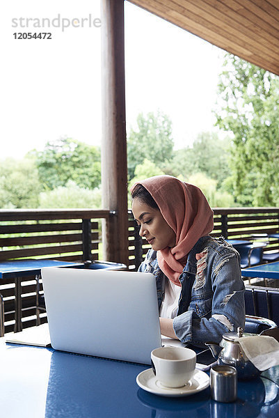Junge Frau  die Hijab trägt und mit ihrem Laptop auf der Terrasse eines Cafés studiert.