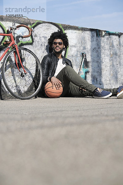 Lächelnder Mann sitzt vor der Grafitti-Wand neben seinem Fahrrad und hält Basketball.