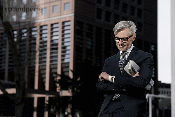 Grauhaariger Geschäftsmann vor rotem Wolkenkratzer mit Blick auf seine smartwatch