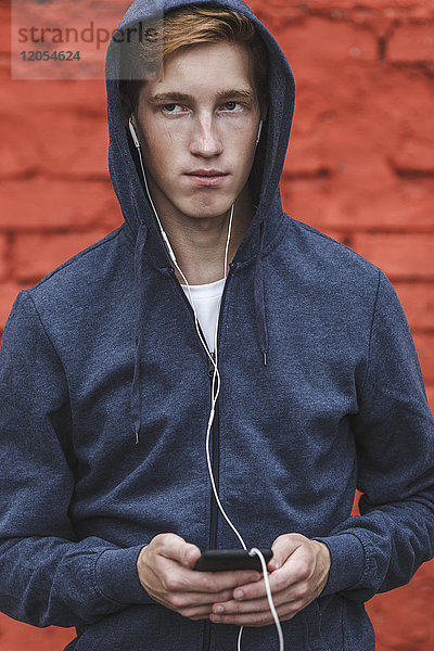 Junger Mann mit Handy und Kopfhörer an roter Ziegelwand