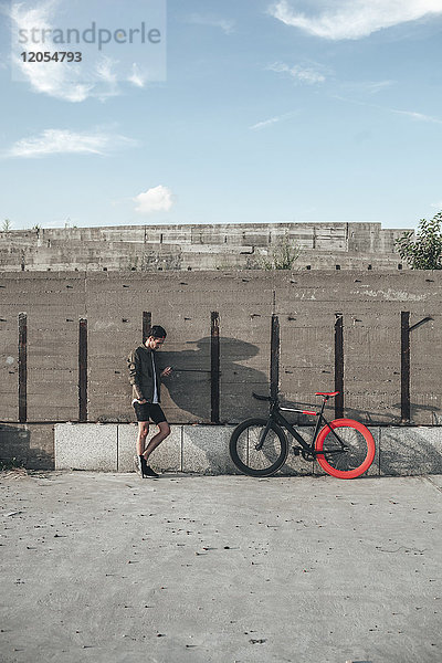 Junger Mann steht an der Betonwand neben dem Fixie Bike mit dem Handy