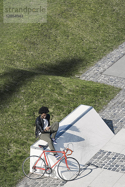 Mann sitzt im Stadtskatepark mit Smartphone neben seinem Fahrrad