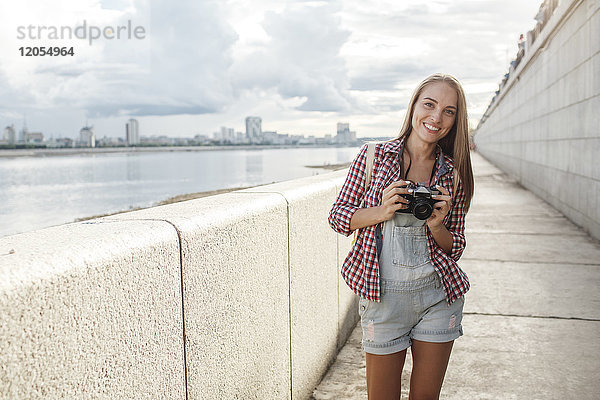 Lächelnde junge Frau mit Kamera am Flussufer