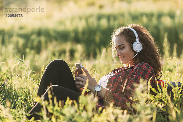 Teenagermädchen hört Musik mit Kopfhörern auf einer Wiese und schaut aufs Handy.