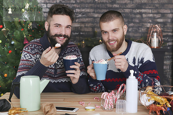 Glückliches schwules Paar trinkt heiße Schokolade mit Sahne und gehackten Zuckerstangen zur Weihnachtszeit.