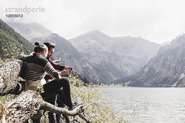 Österreich  Tirol  Alpen  Paar auf Baumstamm am Bergsee mit dem Handy entspannen